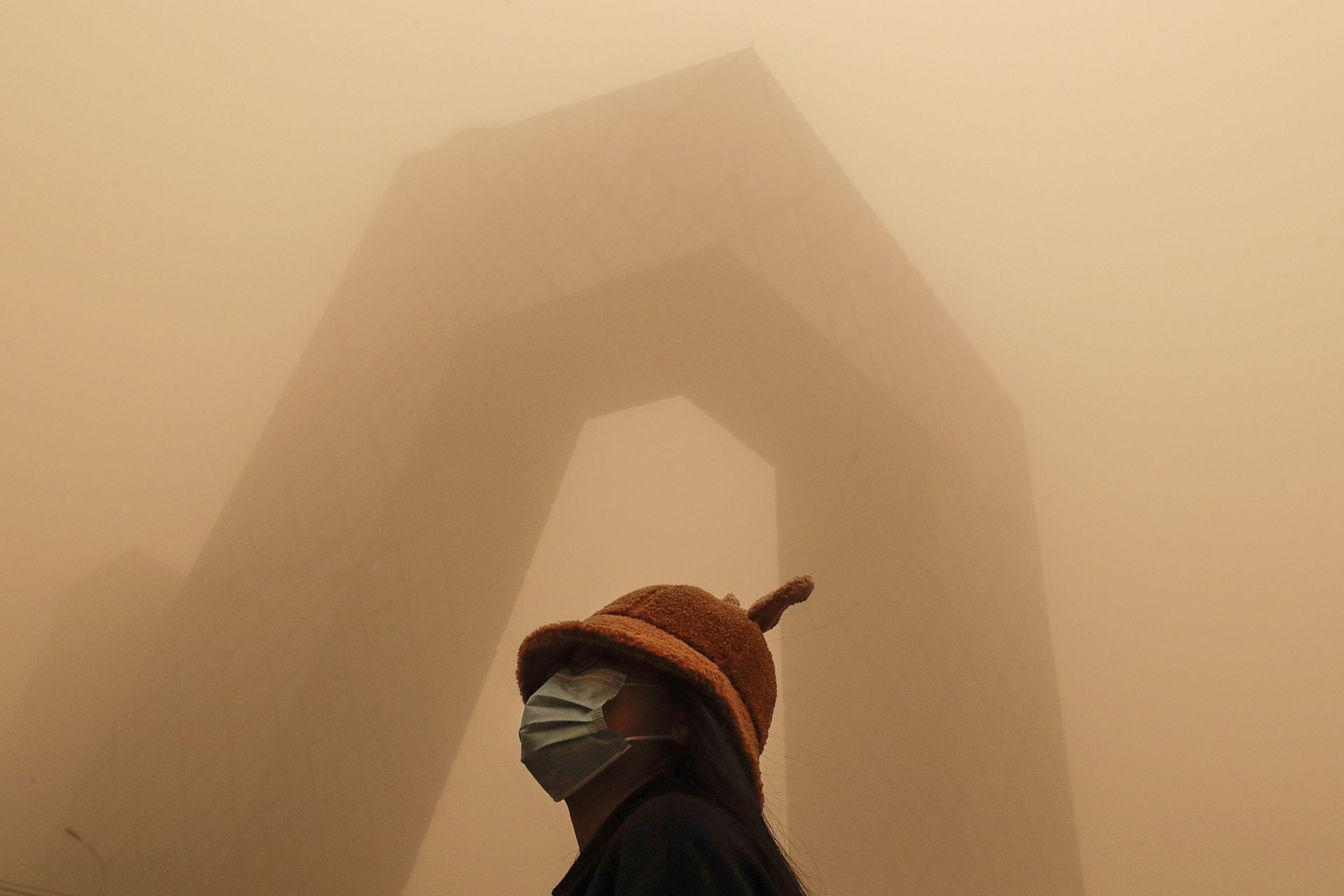 北京受到沙尘暴的袭击，一名戴着口罩的女子站在中国中央电视台大楼对面。（AP）